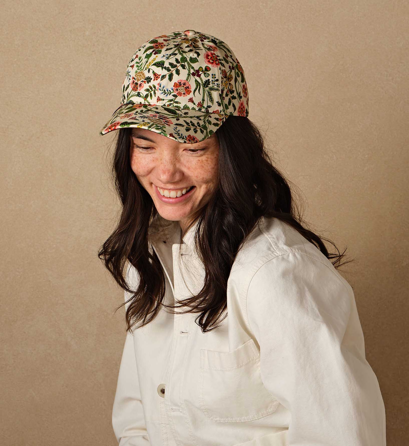 A woman with long dark hair and freckles smiles while looking down, wearing Faire's Gracie Baseball Cap—a floral print, adjustable hat—and a cream button-up shirt against a beige background.