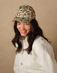 A woman with long dark hair and freckles smiles while looking down, wearing Faire's Gracie Baseball Cap—a floral print, adjustable hat—and a cream button-up shirt against a beige background.