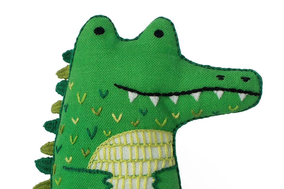The Faire Alligator Embroidery Kit features a green fabric crocodile plush with embroidered eyes, teeth, and colorful patterns inspired by alligator designs, displayed against a white background.