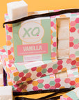 Boxes of Faire Vanilla Marshmallows, with hexagonal pastel designs, are stacked together. Loose vanilla marshmallows and Madagascar vanilla beans surround the allergy-friendly packaging.