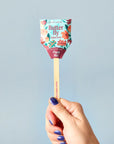 A hand with blue-painted nails holds a Faire Garden Seed Pop featuring a floral wrapper and “Plant Me!” on the stick—ideal for starting seeds in your vegetable garden—set against a light blue background.