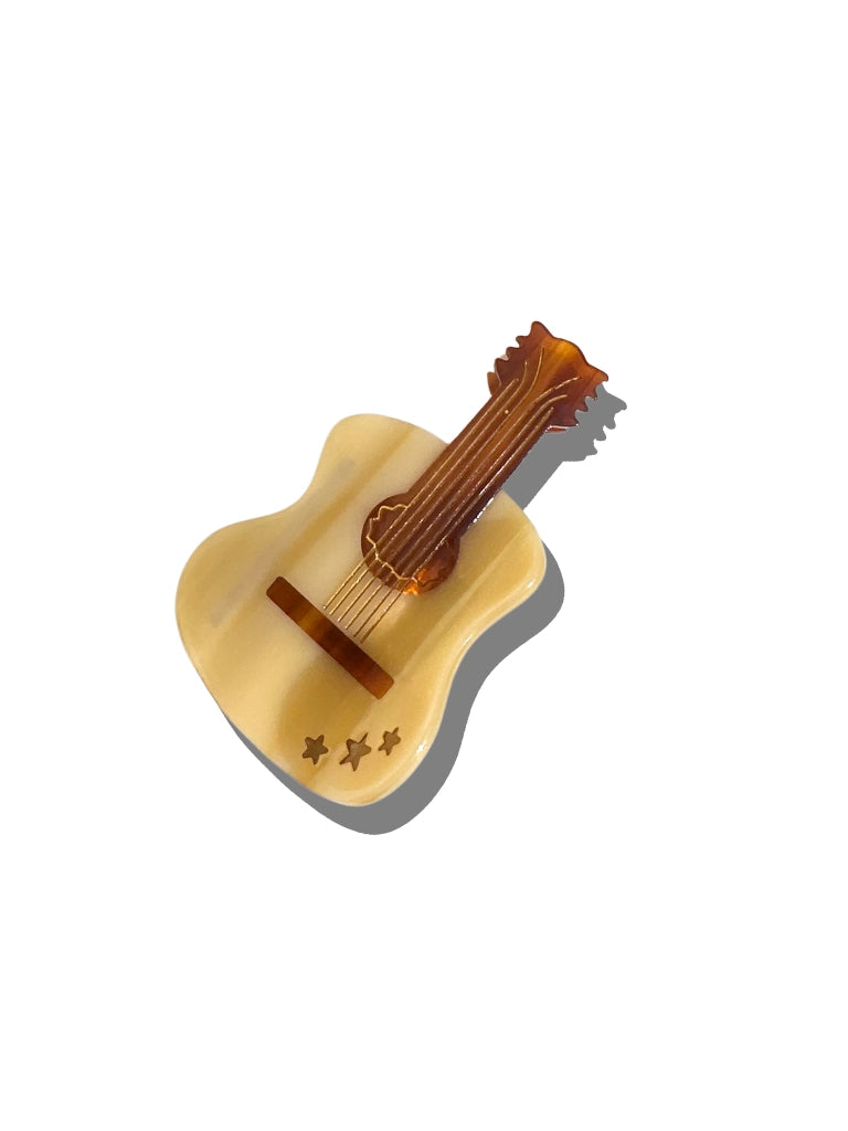 The Faire Fashion Hair Clip is a small, decorative accessory shaped like a guitar, made from eco-friendly cellulose acetate with a cream body, brown neck, and three star cutouts. Shown on a white background with a gray shadow.