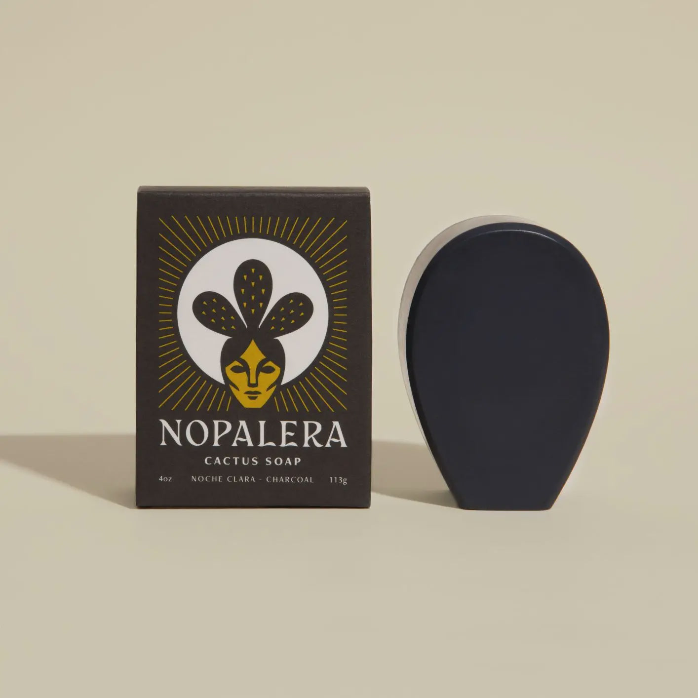 A dark, oval-shaped cacti soap rests beside its packaging, adorned with a stylized face featuring cactus-like hair and the text "Faire Noche Clara Cactus Soap, Charcoal infused with Prickly Pear Cactus, 113g," set against a light beige backdrop.