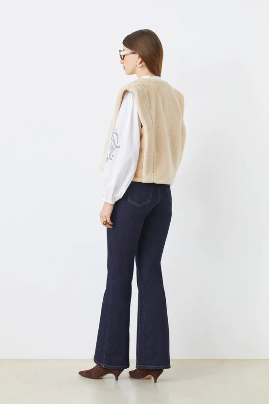 A woman with straight brown hair faces away, wearing SUNCOO's Diego Vest in beige recycled polyester faux fur over a white long-sleeve blouse, dark flared jeans, and brown pointed-toe shoes. She completes the look with sunglasses.