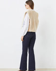 A woman with straight brown hair faces away, wearing SUNCOO's Diego Vest in beige recycled polyester faux fur over a white long-sleeve blouse, dark flared jeans, and brown pointed-toe shoes. She completes the look with sunglasses.
