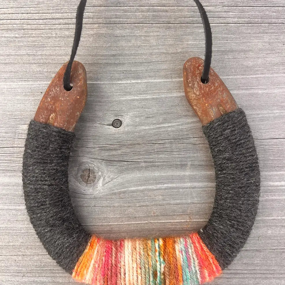 The Lucky Horseshoe by Faire features upcycled horseshoes wrapped in dark gray and multicolored yarn with orange, pink, yellow, and teal accents. Hung by a black cord, this unique piece makes a personalized gift.