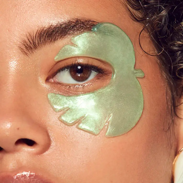 Close-up of a person with curly hair, a green tropical leaf-shaped gel patch from Faire's Cucumber/Tea Eye Mask is applied under their right eye. Their skin looks dewy and moisturized.