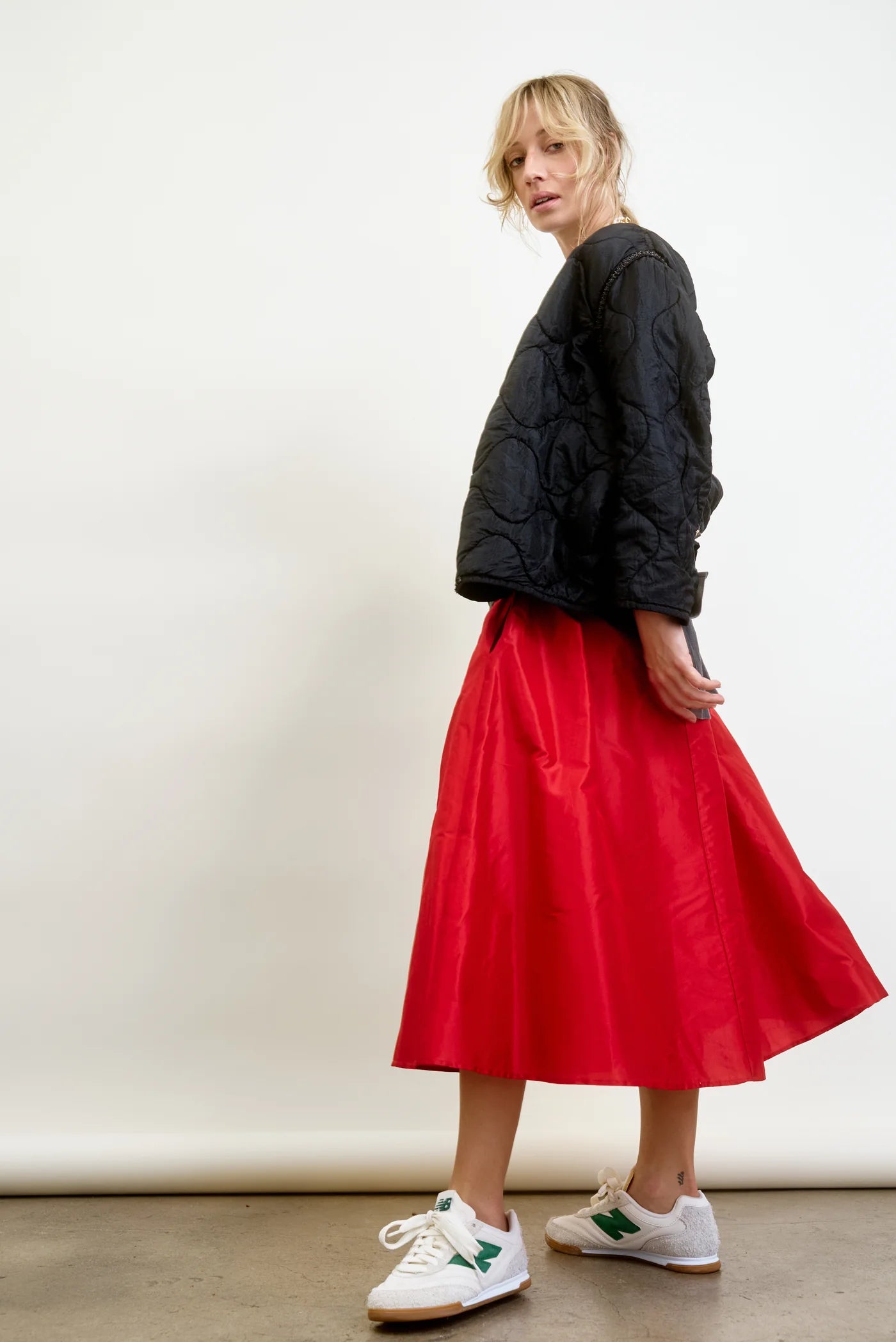 Dressed in a black quilted jacket and the red LE SKIRT Silk Taffeta by Aquarius Cocktail, a person exudes elegance. White sneakers with green accents lend a modern 40's flair to their look, as they pose stylishly against a minimalistic white backdrop.