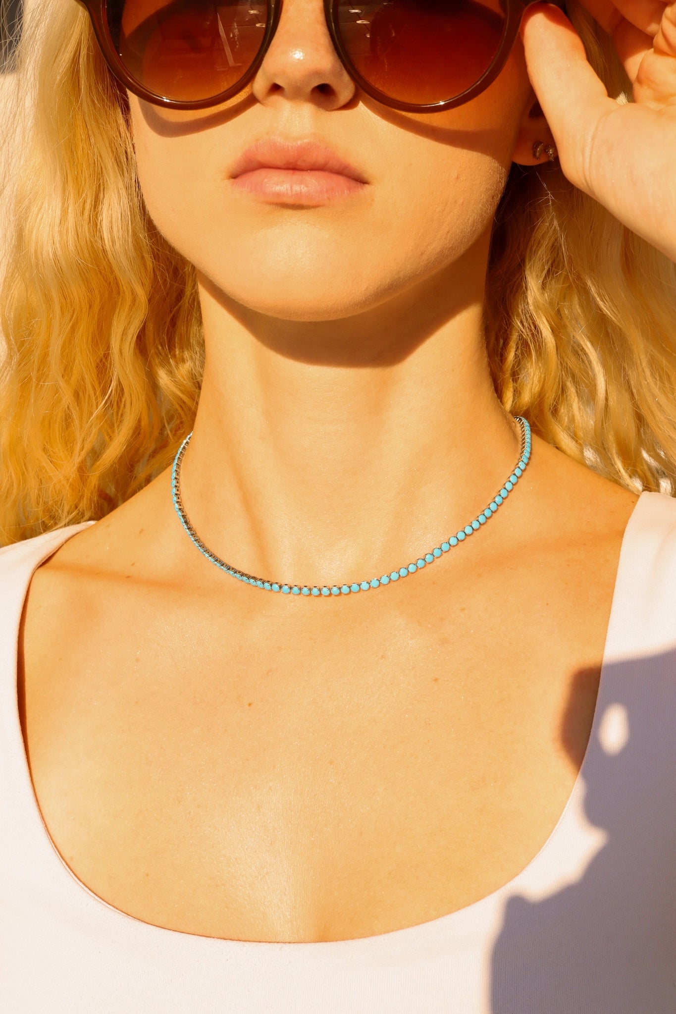 A woman in a white top and large brown sunglasses wears the Faire Turquoise Tennis Necklace, crafted from hypoallergenic 316L stainless steel, featuring sparkling rhinestones, with her wavy blonde hair glowing in sunlight.
