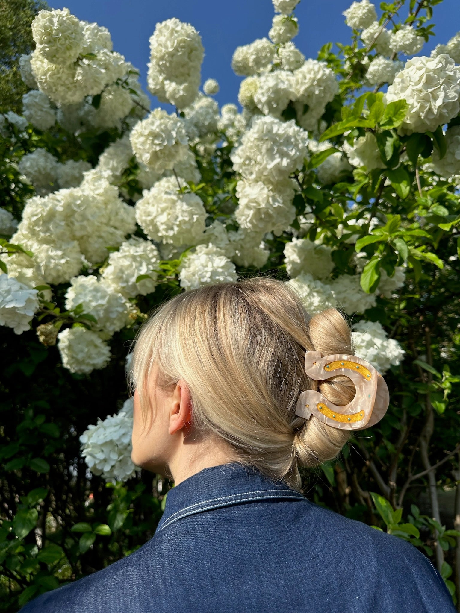 A person with blonde hair wears the Faire Fashion Hair Clip, crafted from eco-friendly cellulose acetate, while standing outdoors in a denim jacket near white flowering bushes under a clear blue sky.