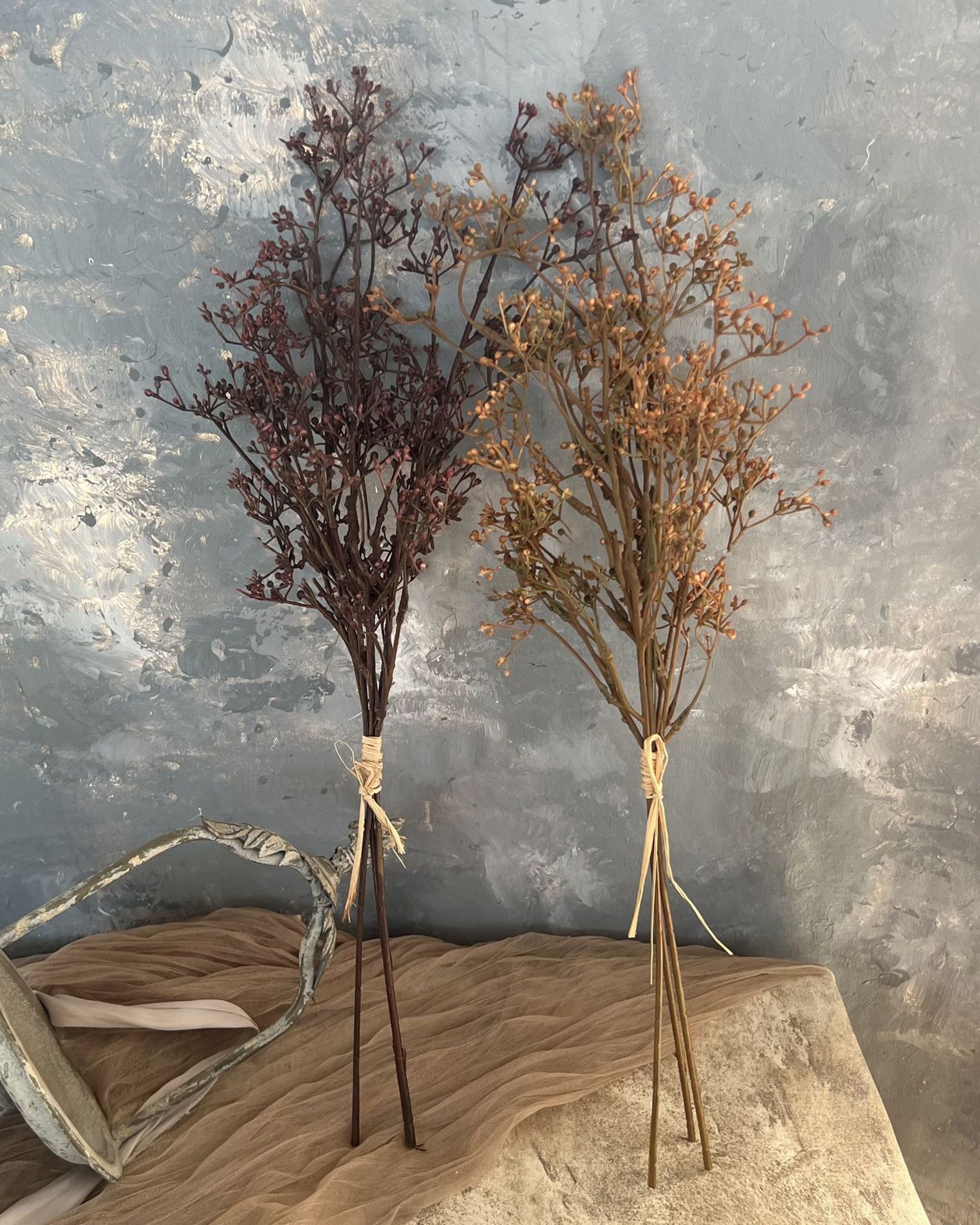 Two small 22" Faux Seeded Baby's Breath Bunches by Faire—one dark reddish-brown, one golden-brown—are tied with twine and set against a textured gray-blue background with a draped tan cloth.