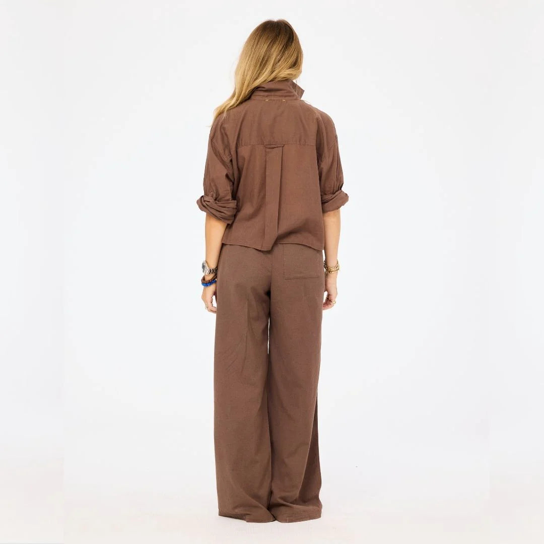 A person with long blonde hair stands facing away in the Kerri Rosenthal Tia Love Brigade Shirt—a relaxed button-down brown top with rolled sleeves—paired with matching wide-leg pants, set against a plain white background.