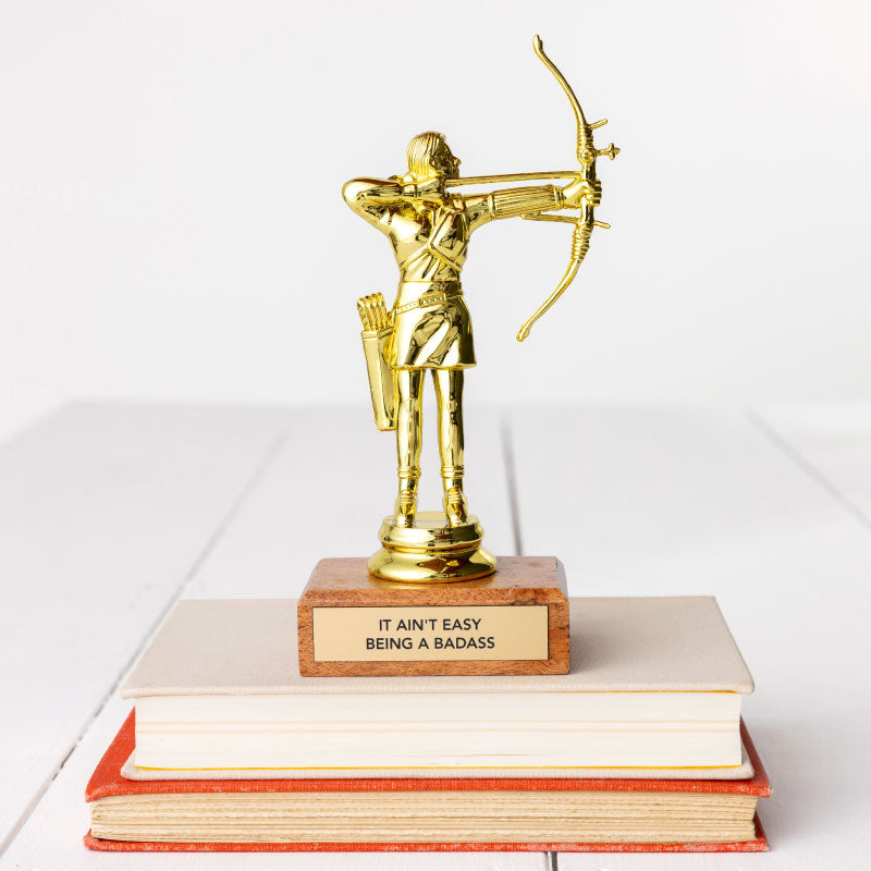 A golden JE Trophy depicting a figure with a bow and arrow, standing on a stack of books in Arizona style. The base of the trophy has a plaque that reads &quot;IT AIN&#39;T EASY BEING A BADASS.&quot; (Faire)