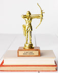 A golden JE Trophy depicting a figure with a bow and arrow, standing on a stack of books in Arizona style. The base of the trophy has a plaque that reads "IT AIN'T EASY BEING A BADASS." (Faire)