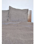 A neatly made bed displays the Modernplum Baltic Queen Duvet in brown and white thin stripes with an invisible zipper, plus a matching pillowcase, set against a wooden headboard and plain white wall.