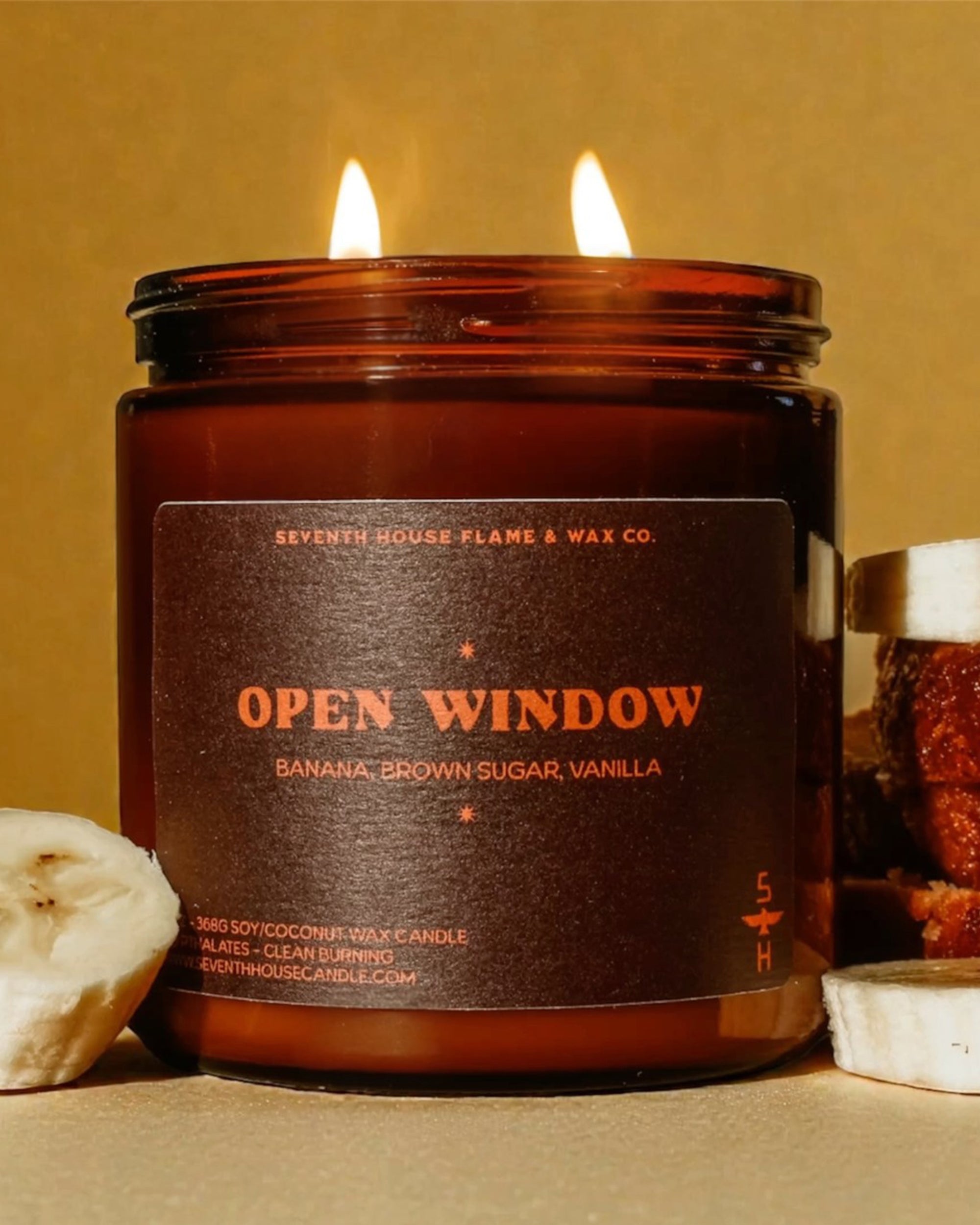 The Faire Banana Nut Bread Candle, labeled "OPEN WINDOW" and featuring banana, brown sugar, and vanilla notes, sits beside banana slices and banana bread on a beige background.