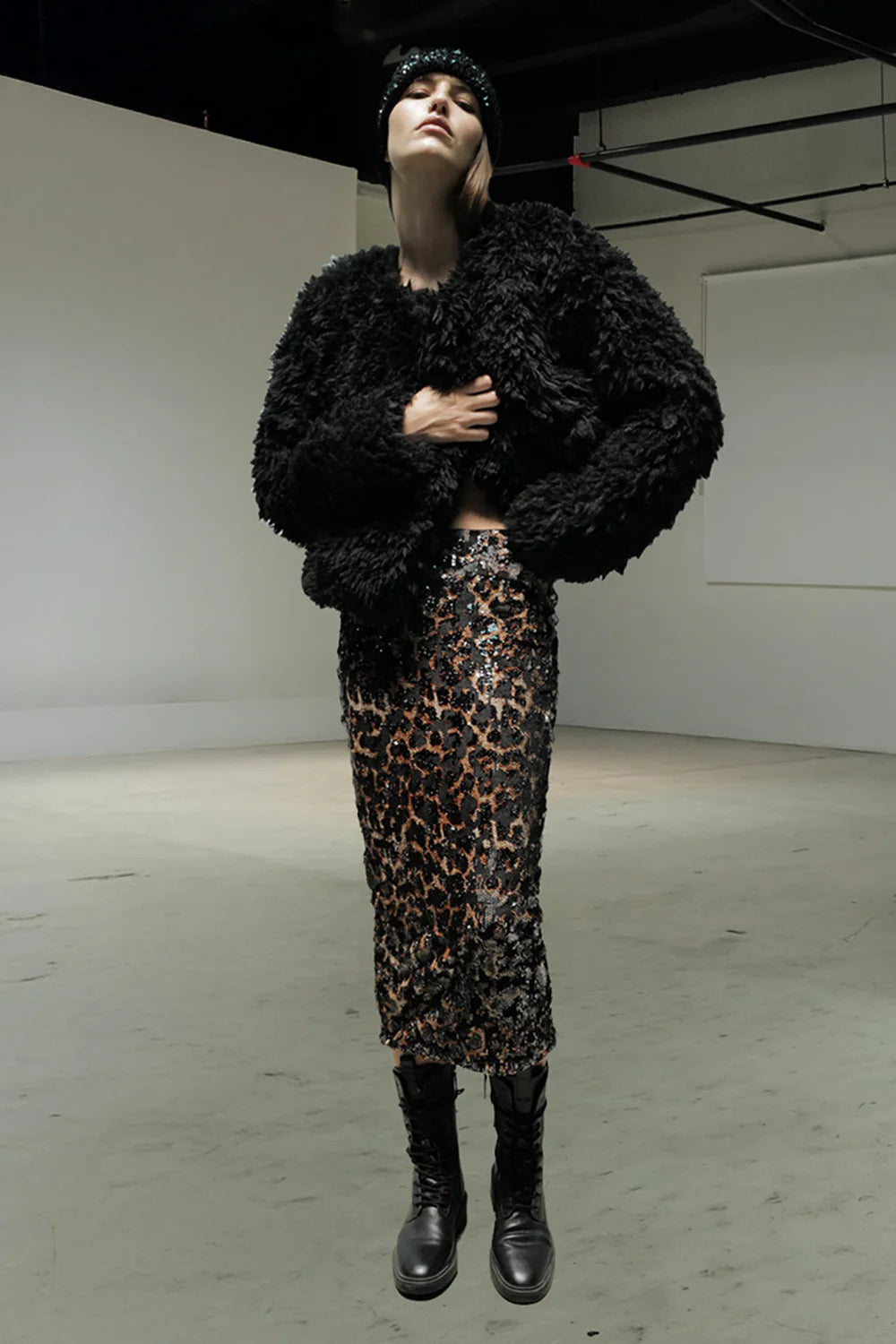 A person wearing a textured black faux fur jacket, the Le Superbe Liza Skirt, black lace-up boots, and a black beanie stands in a minimalist industrial-style space.