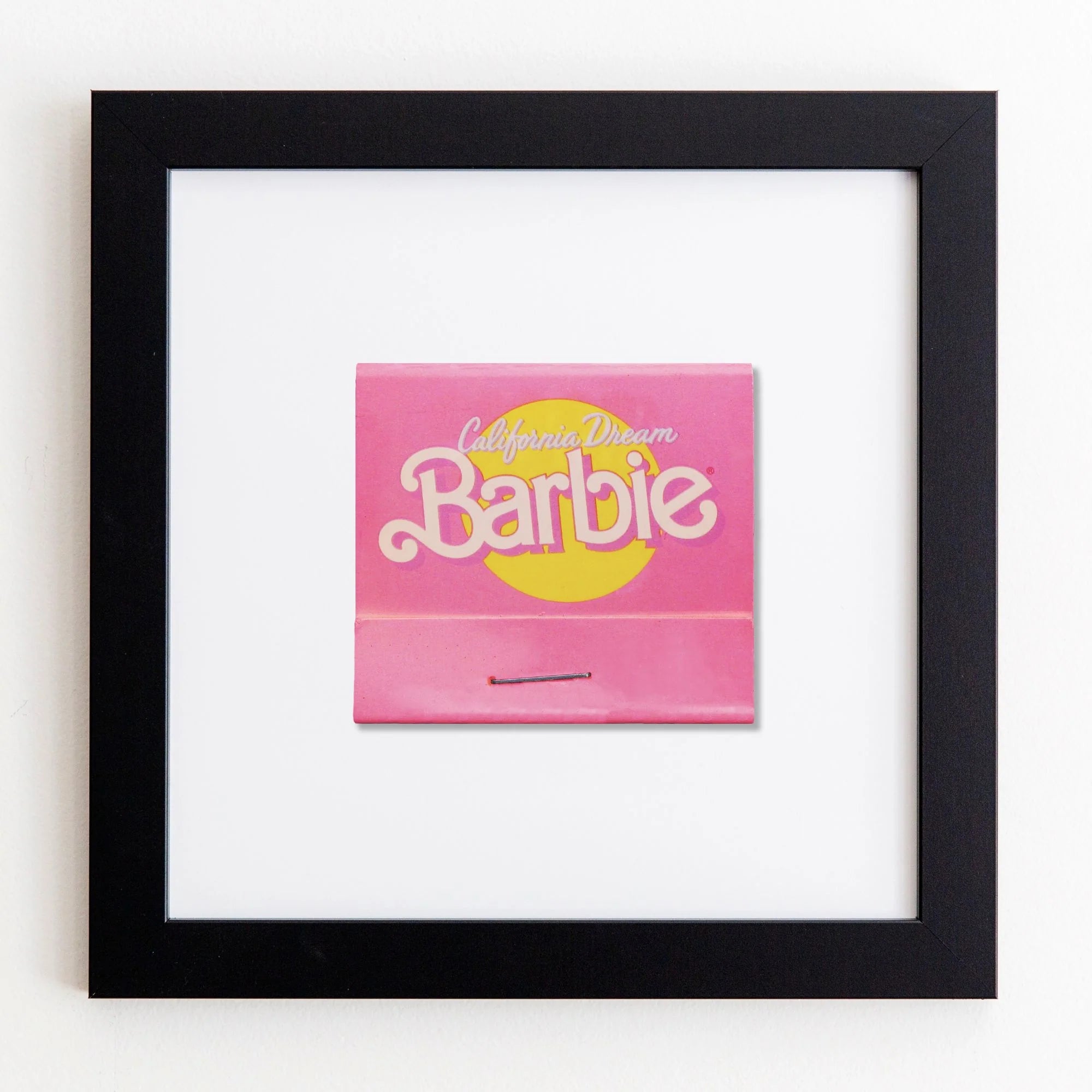 A framed Barbie board game titled &quot;California Dream,&quot; prominently displaying a pink box with yellow accents, mounted on a white acrylic background within a Match South Art Square Black Frame.