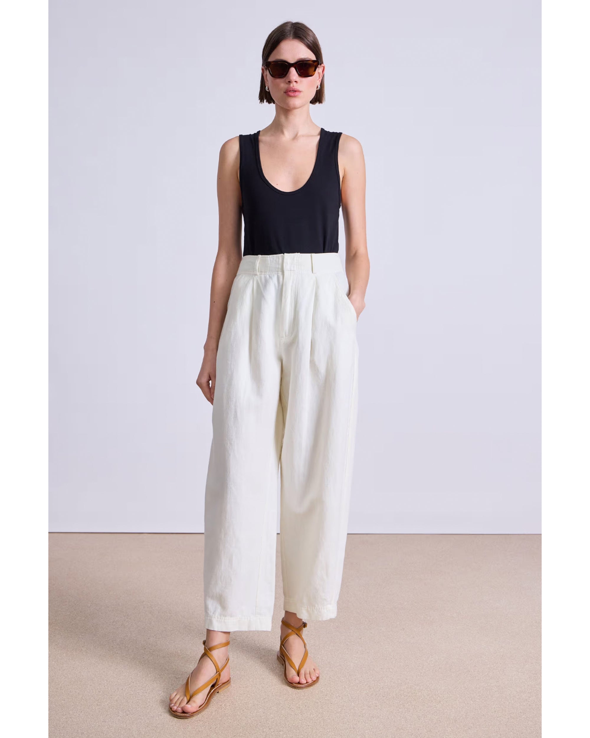 A woman stands against a plain background wearing sunglasses, a sleeveless black top, and Apiece Apart's Bari Crop Trouser. She completes her look with tan strappy sandals and keeps her hands in her pockets.