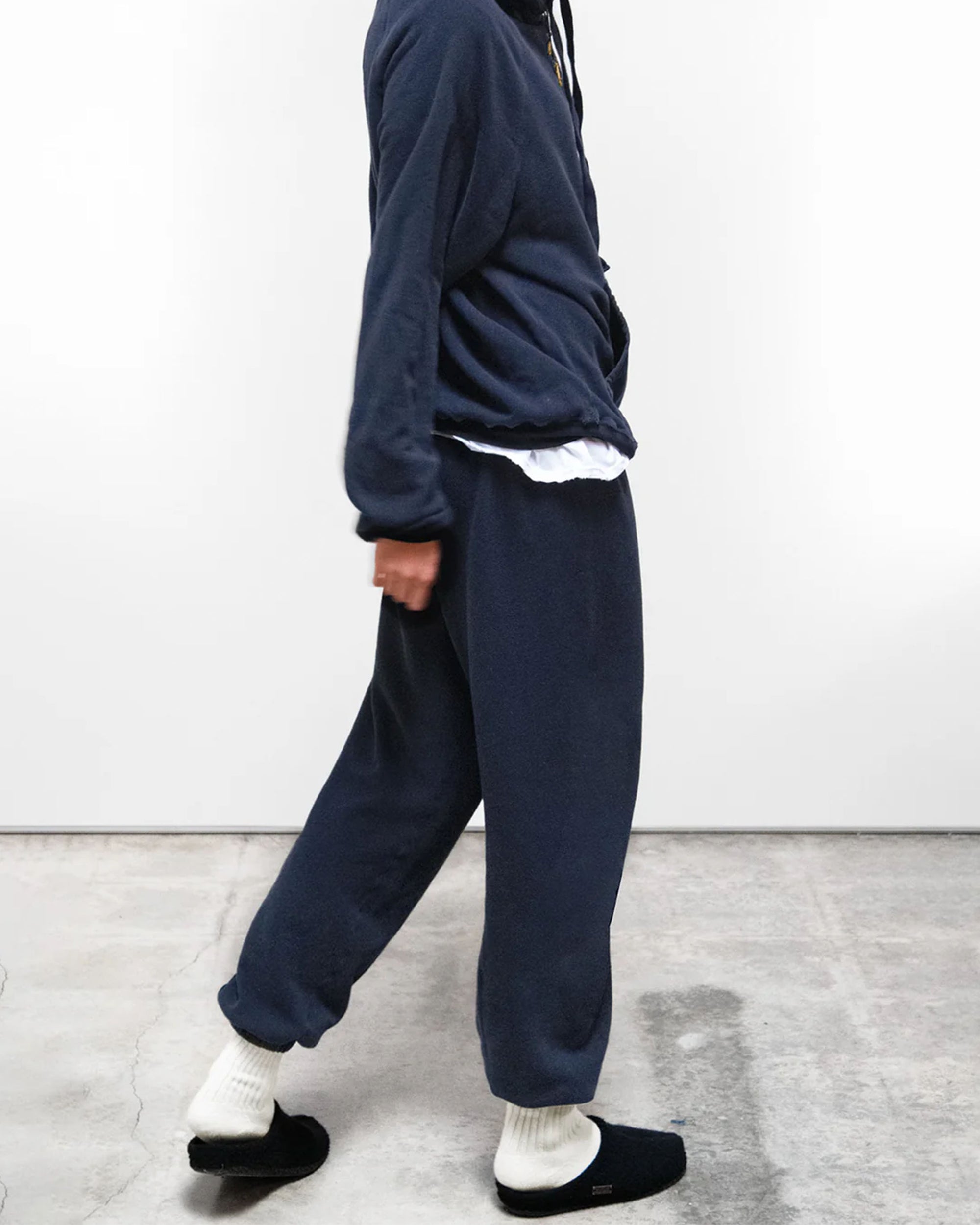 A person indoors wears Free City (sparrow, LLC) BASIC GOODNESS OG SWEATPANTS—loose-fitting, navy blue, unisex French terry sweatpants made in the USA—with a matching sweatshirt, white undershirt, white socks, and black slip-on shoes. Face not shown.