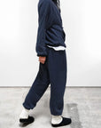 A person indoors wears Free City (sparrow, LLC) BASIC GOODNESS OG SWEATPANTS—loose-fitting, navy blue, unisex French terry sweatpants made in the USA—with a matching sweatshirt, white undershirt, white socks, and black slip-on shoes. Face not shown.