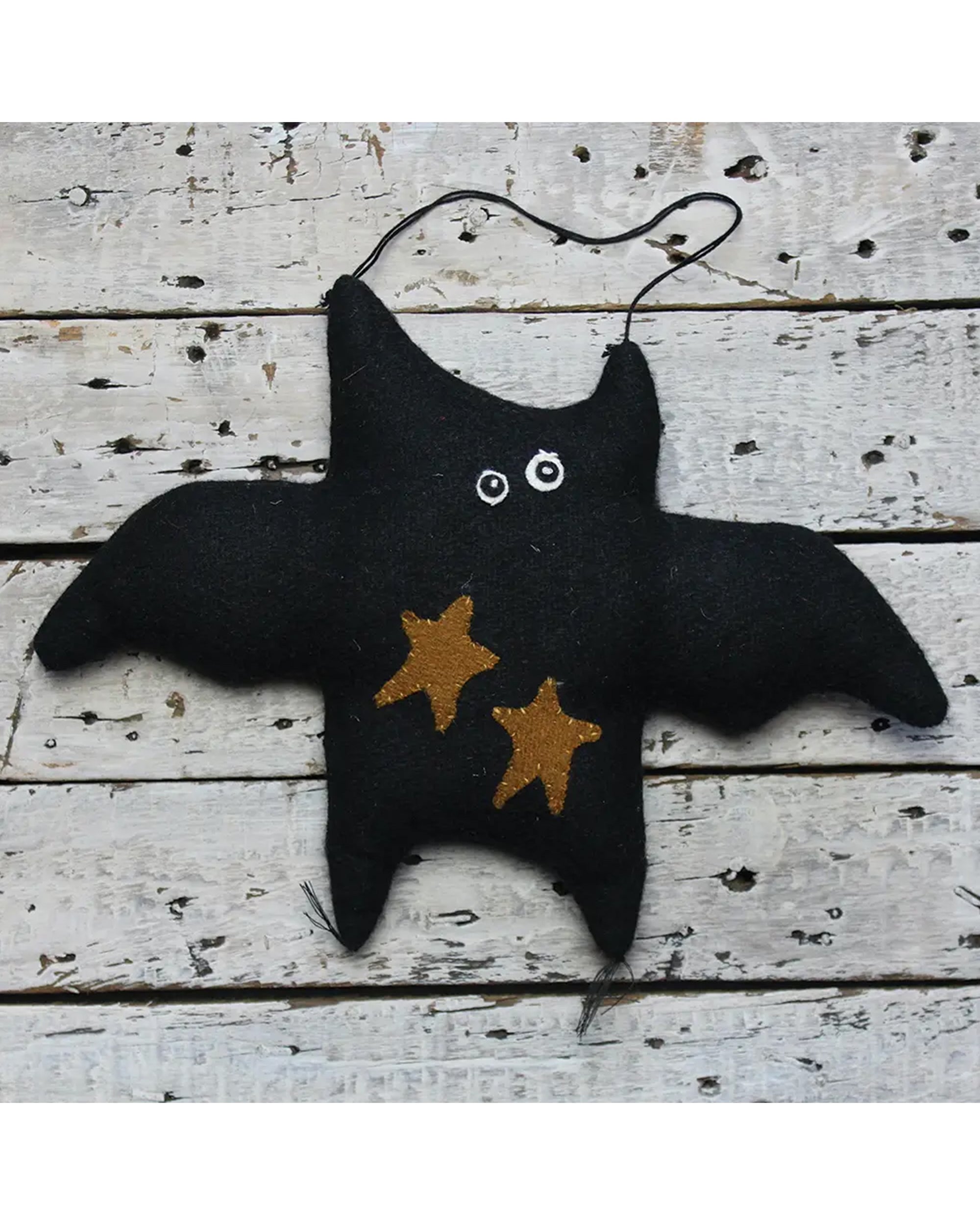 The Faire Bat Door Hanger is a handmade black felt ornament with gold stars and googly eyes, shown on a distressed white wood surface—ideal rustic or farmhouse fall decor.