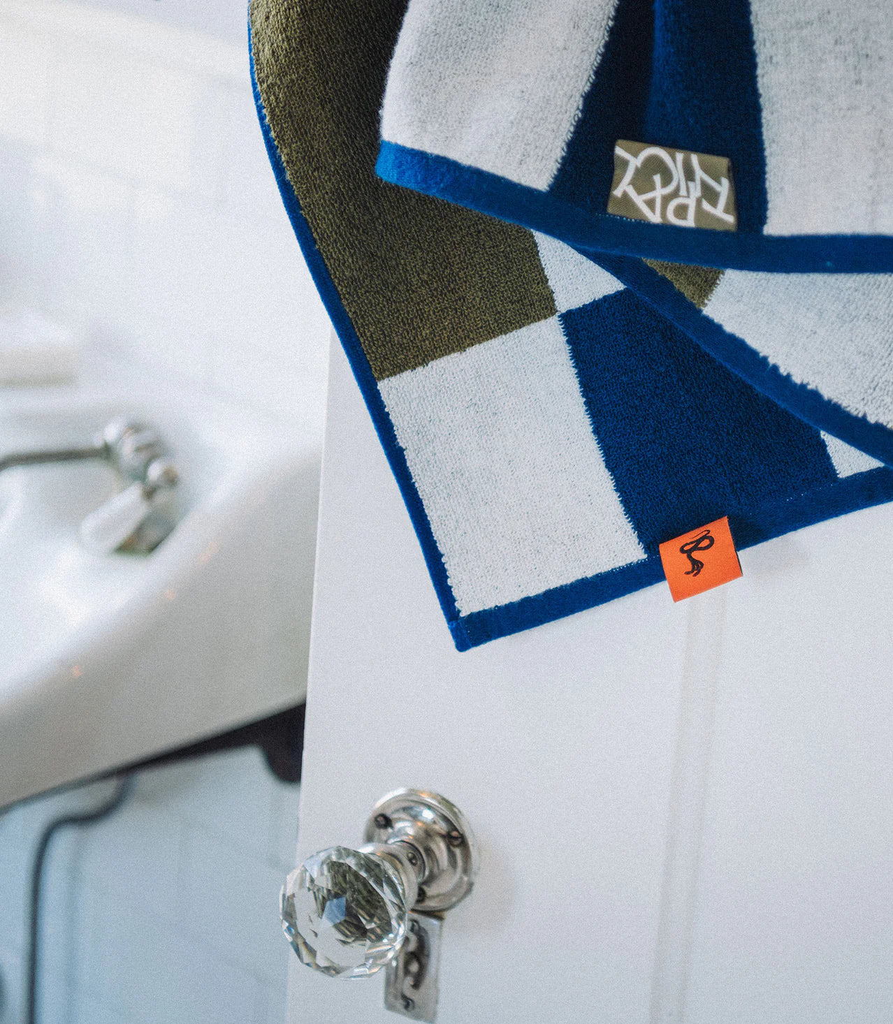 The Typical Navy/Moss Stripe Bath Towel, featuring blue, white, and olive stripes with an orange tag, hangs over a white door with a glass knob; part of a white sink appears in the background.