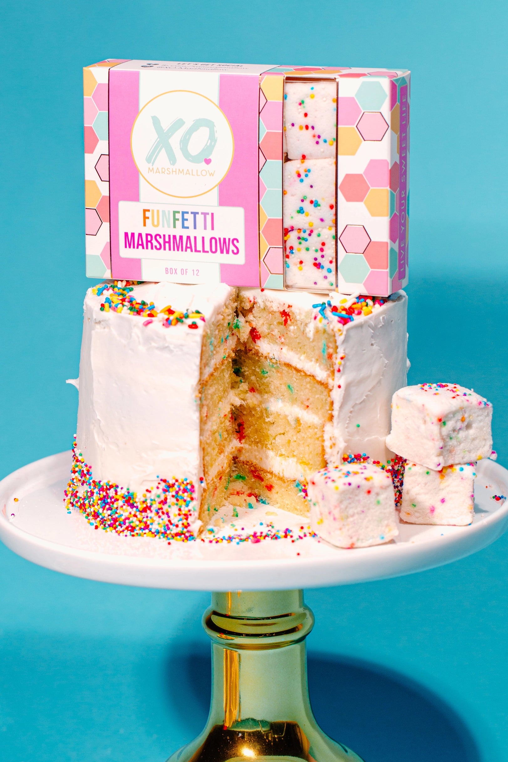 A white frosted funfetti cake with rainbow sprinkles sits on a gold stand, with slices and cubes cut out. Behind it is a pastel box of Faire Funfetti Marshmallows, which are allergy-friendly.