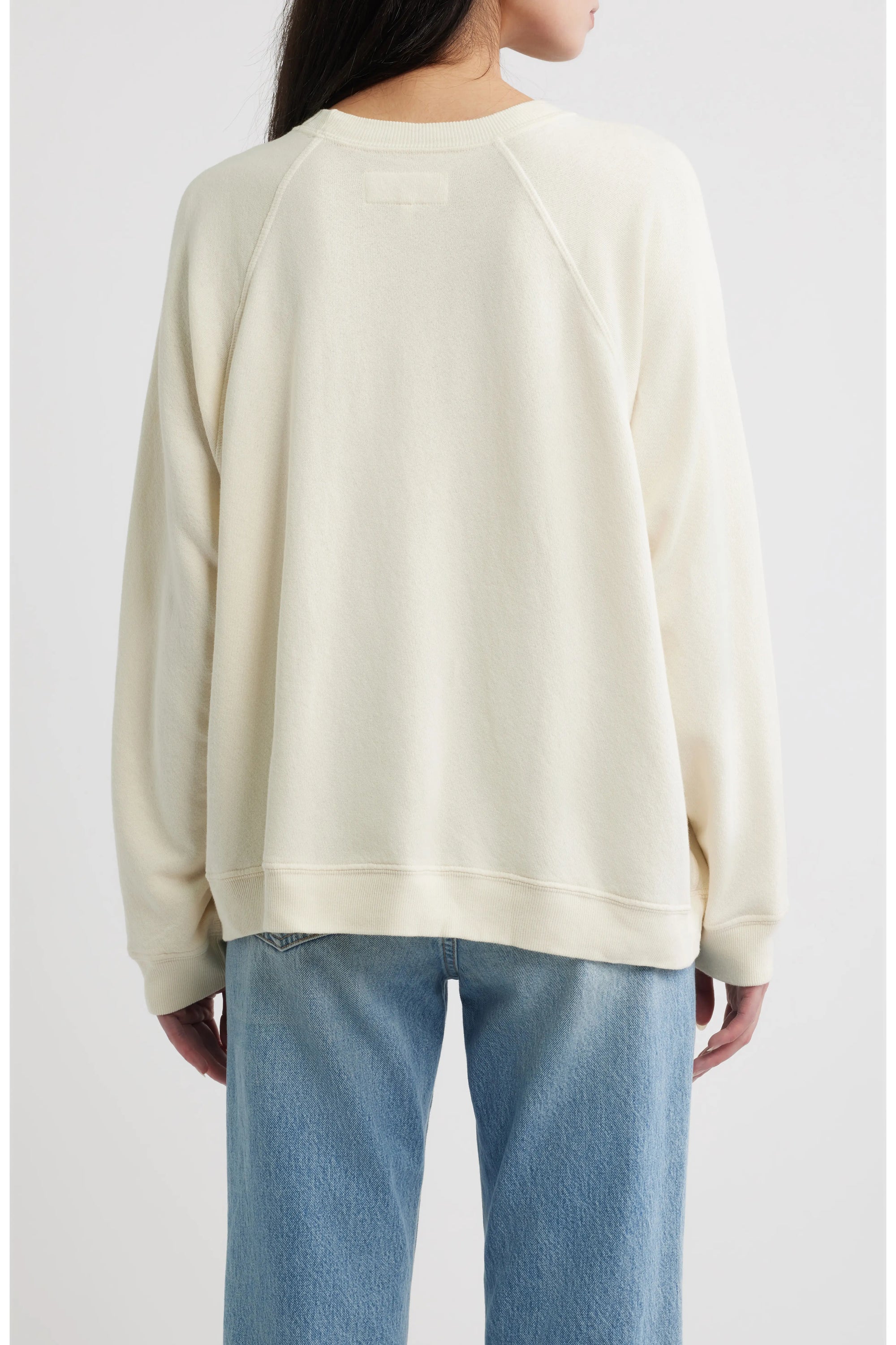 A person is shown from the back against a plain white background, wearing The Great Inc.'s The Rehearsal Sweatshirt in cream-colored cotton paired with light blue jeans.