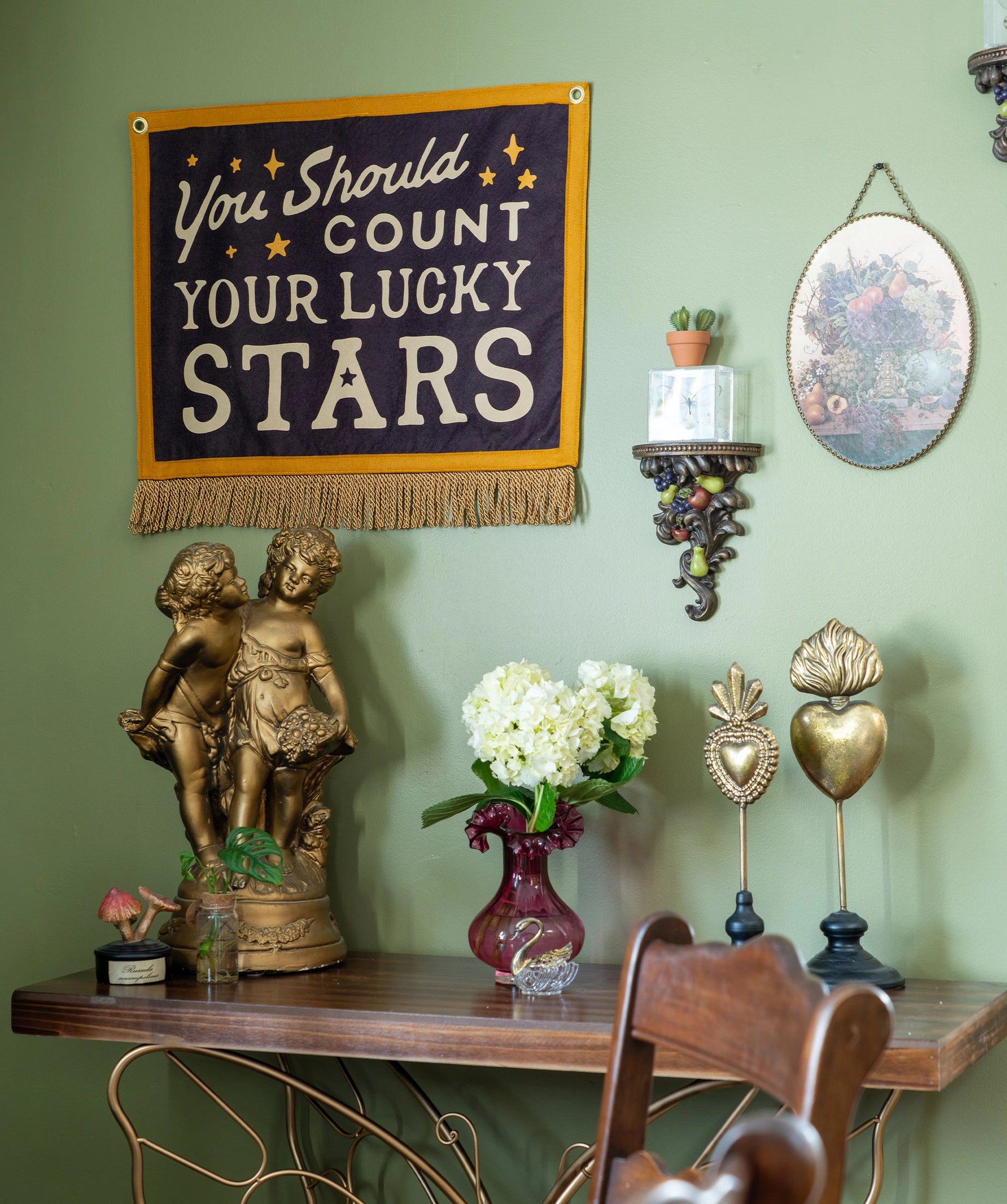 A decorative vignette showcases Faire's Count Your Lucky Stars Camp Flag handmade wall decor, a vase of white flowers, gold figurines, a framed floral print, and a small potted plant on a light green wall above a wooden table.