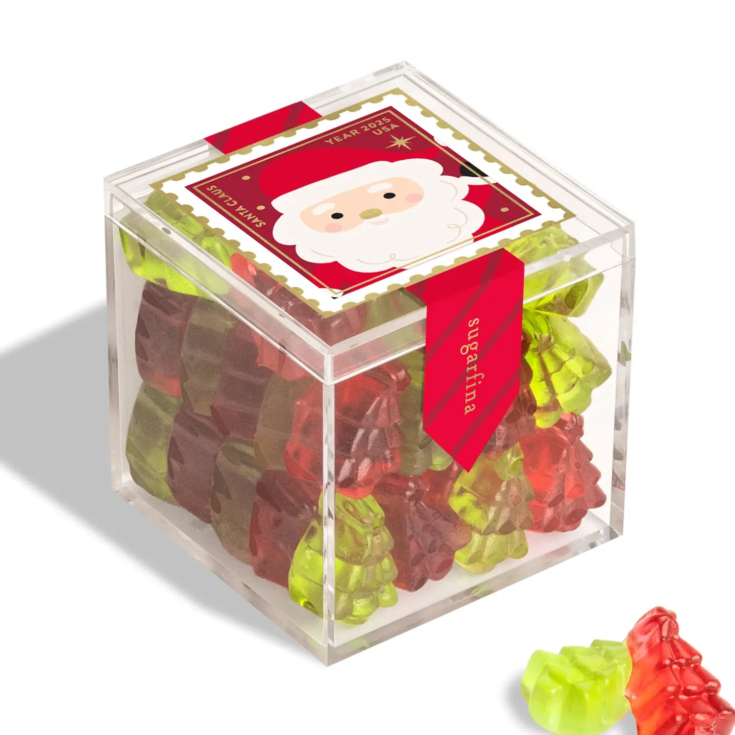 A clear plastic Christmas Candy Cube by Faire, filled with red and green tree-shaped candies, features a festive Santa label on top and a red "sugarfina" ribbon on the side. Two gummies are displayed outside the box.