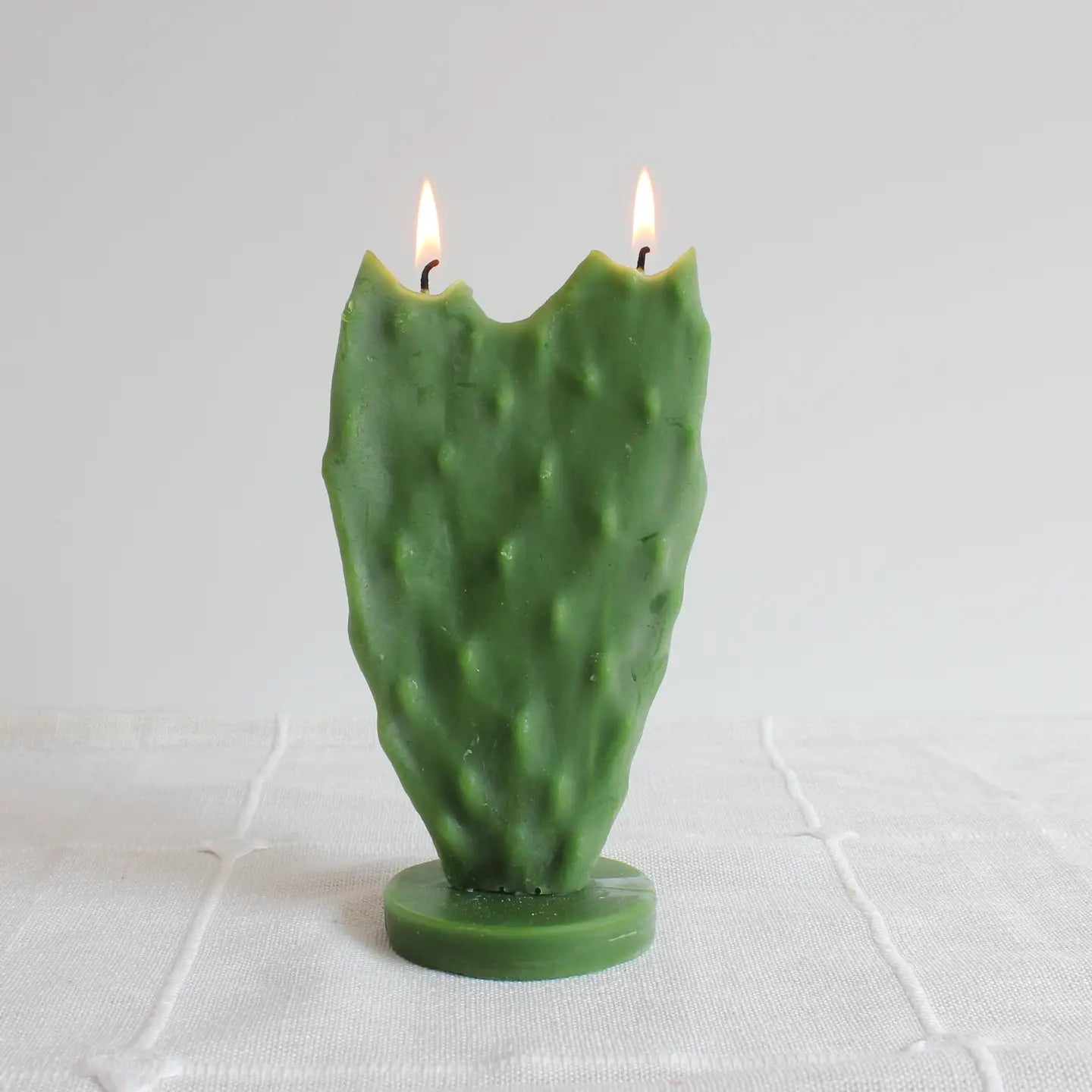 The Nopal Cactus Beeswax Candle by Faire features a green, paddle-shaped cactus design with dual flames, standing upright on a round base—a striking home décor piece shown on a white textured cloth with a plain background.