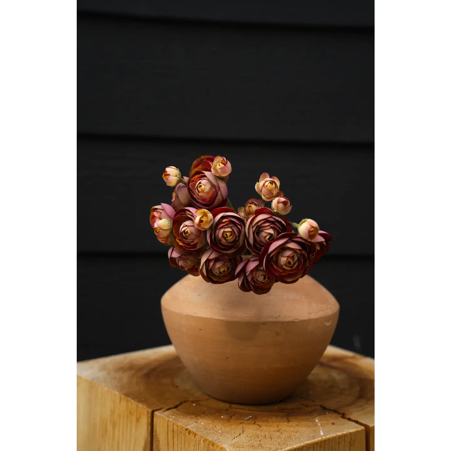 The Indaba Ranunculus Bouquet with vibrant red and yellow blooms is displayed in a round terracotta vase on a wooden surface, set against a dark background.
