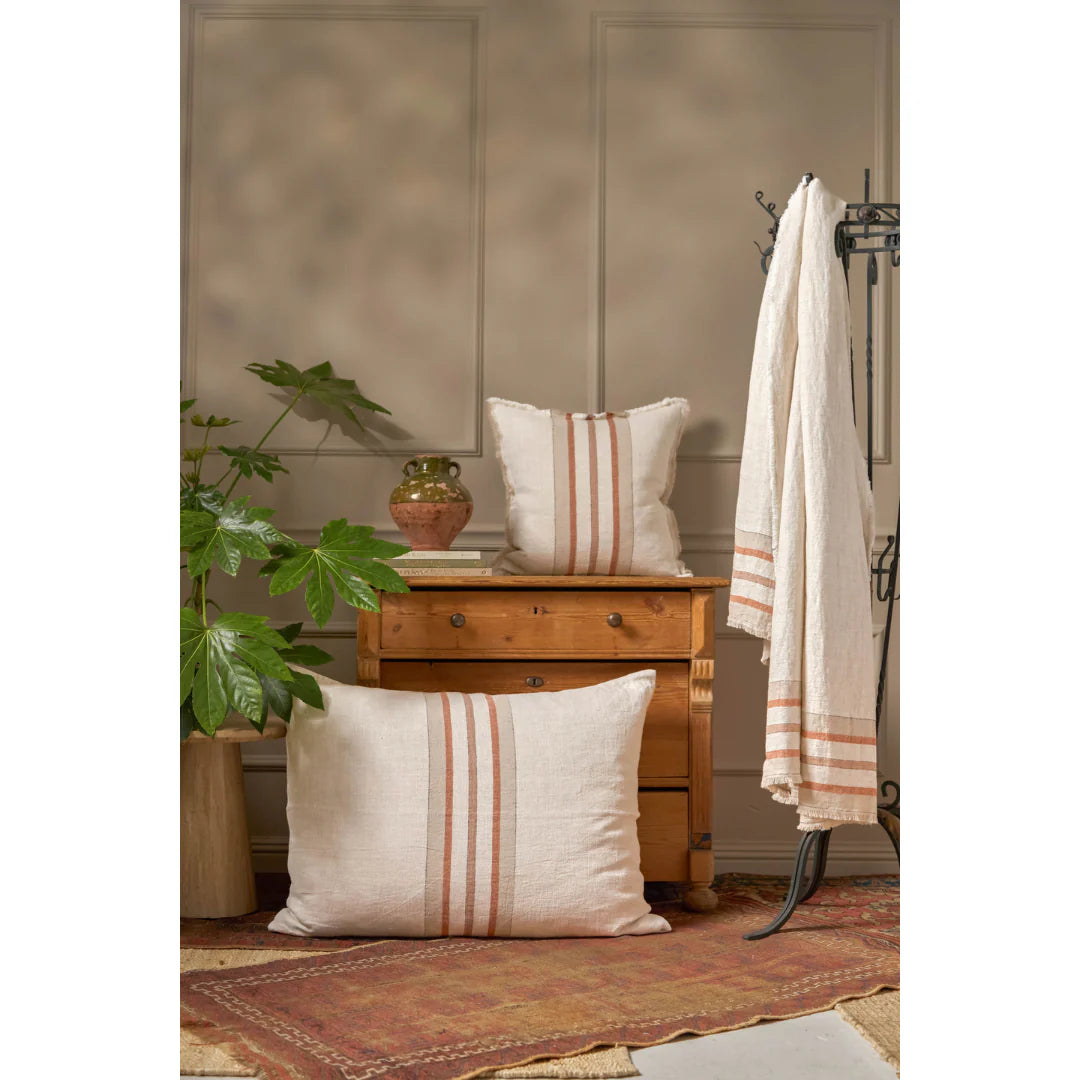 A cozy room featuring a small wooden chest, two POM POM Beck Big Pillows 28x36 in soft linen with rust-colored stripes, a matching throw on a coat rack, a large green plant, a clay vase, and a patterned rug on the floor.