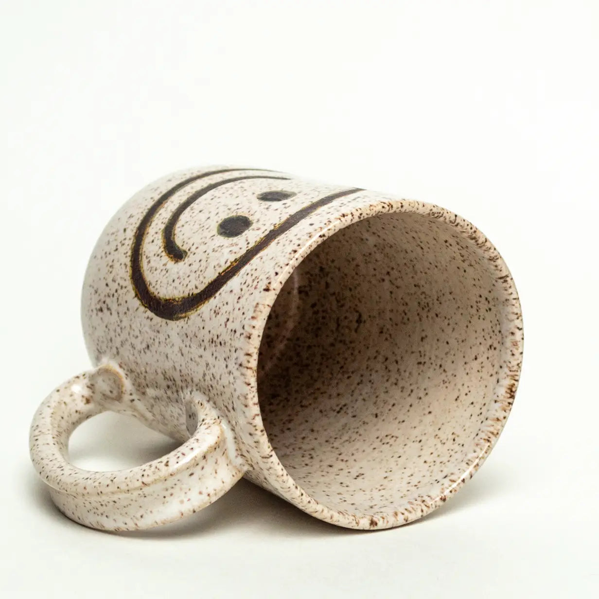 A Faire Ceramic Mug in speckled ivory with a black smiley face, handle left, rests on its side against a plain white background—one of our charming handmade mugs.