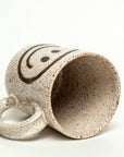 A Faire Ceramic Mug in speckled ivory with a black smiley face, handle left, rests on its side against a plain white background—one of our charming handmade mugs.