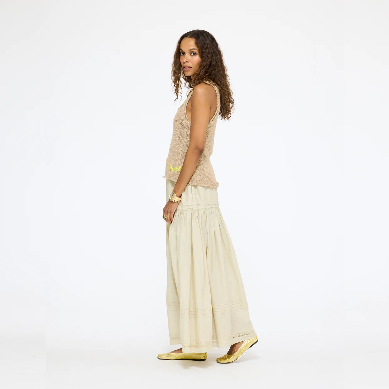 A woman with curly hair stands sideways against a plain white background, wearing a sleeveless beige top, gold flat shoes, and the cream-colored Kerri Rosenthal Vacance Pintuck Skirt.