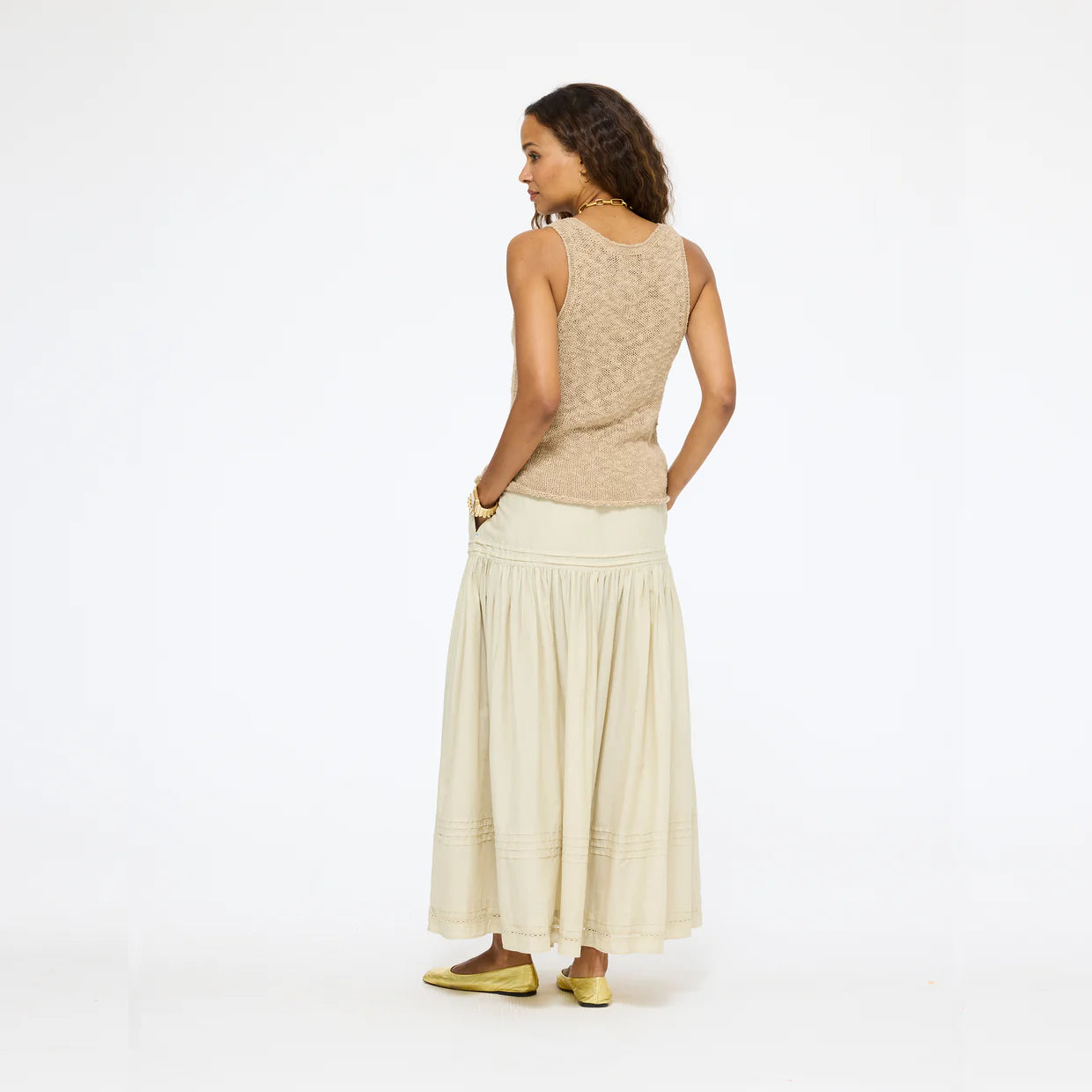 A woman faces sideways with her head turned, wearing a beige sleeveless top, gold flats, and the Kerri Rosenthal Vacance Pintuck Skirt with tiered lace trim, all set against a plain white background.
