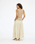A woman faces sideways with her head turned, wearing a beige sleeveless top, gold flats, and the Kerri Rosenthal Vacance Pintuck Skirt with tiered lace trim, all set against a plain white background.