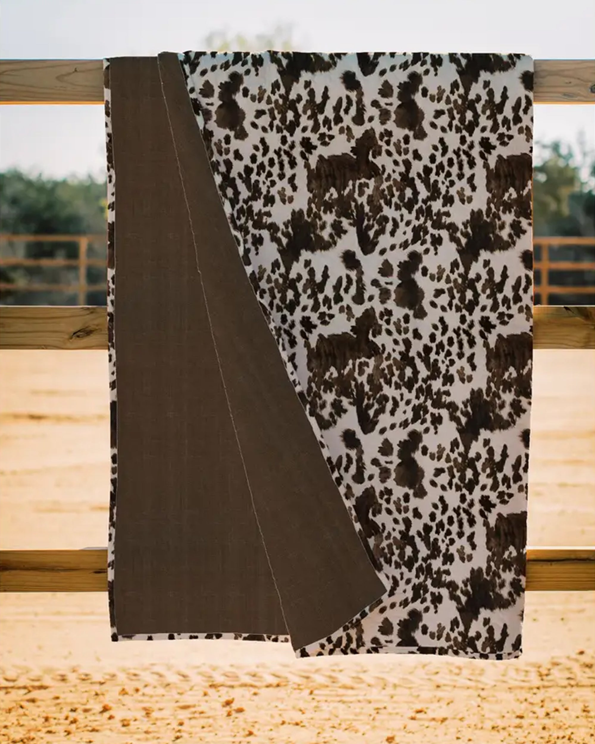 The Faire Bella Hide Brown Quilt, featuring a brown and white cowhide pattern with a solid brown underside, hangs over a wooden fence outdoors, bringing Fort Worth Texas style to any setting.