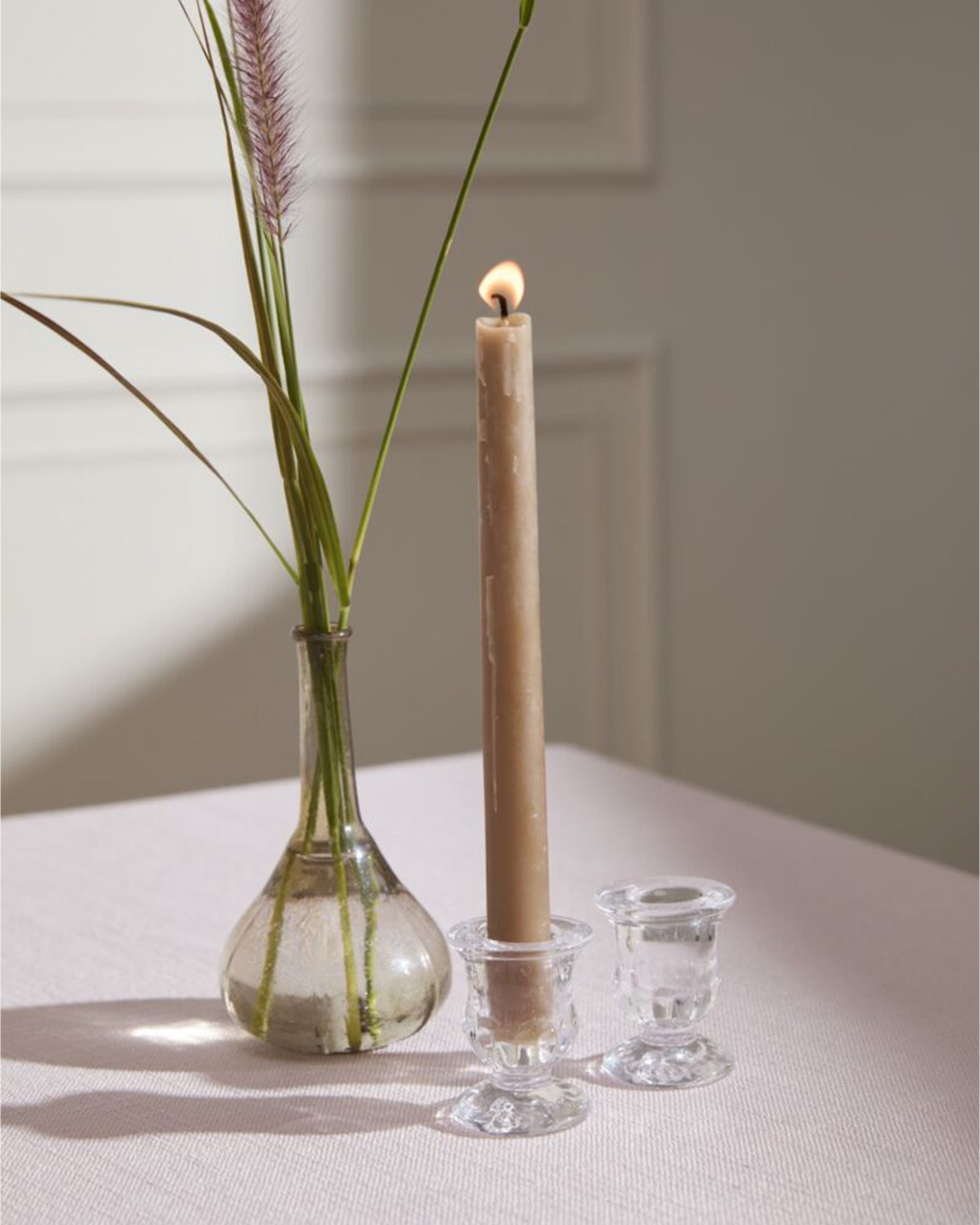 A beige candle glows in the Accent Decor Bellamy Candleholder atop a light table near a vase with green stems and purple grass—an elegant accent for refined events. Soft sunlight adds gentle shadows.