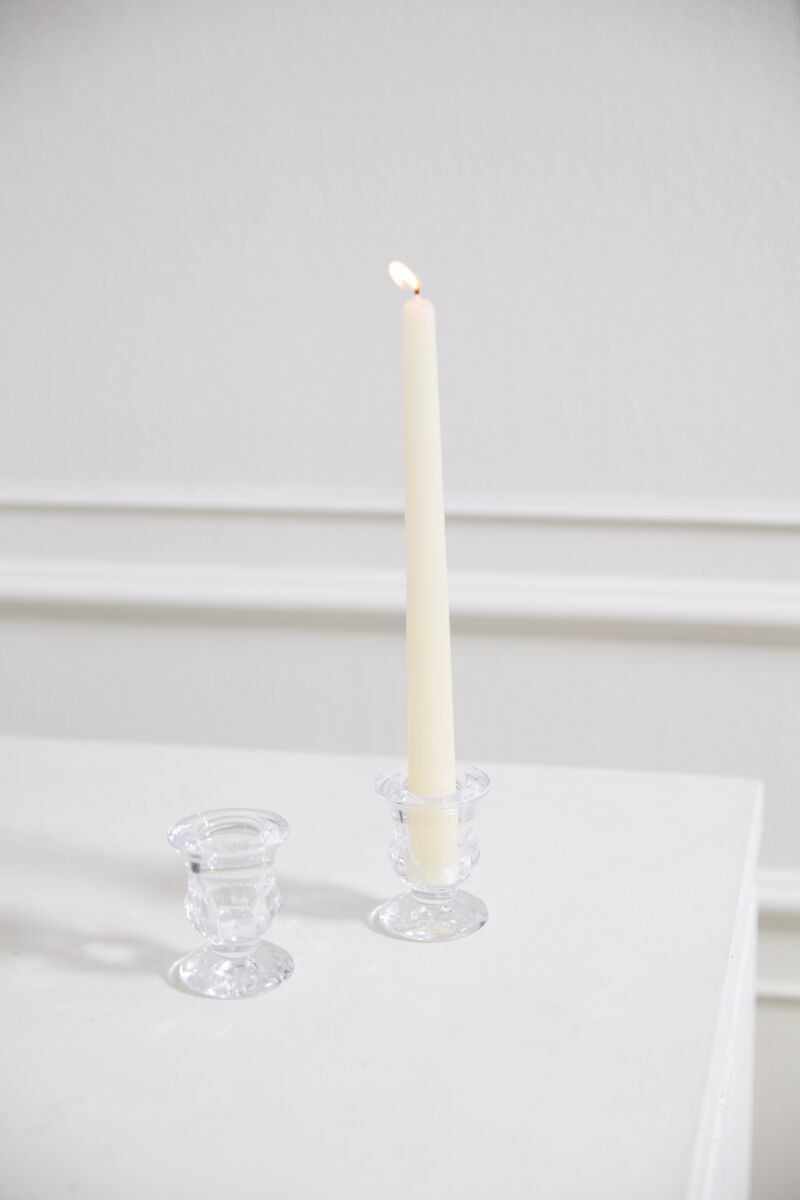 A tall, lit white candle in an urn-shaped clear Bellamy Candleholder by Accent Decor sits on a white surface beside an empty matching holder, all set against a white background with decorative wall molding.