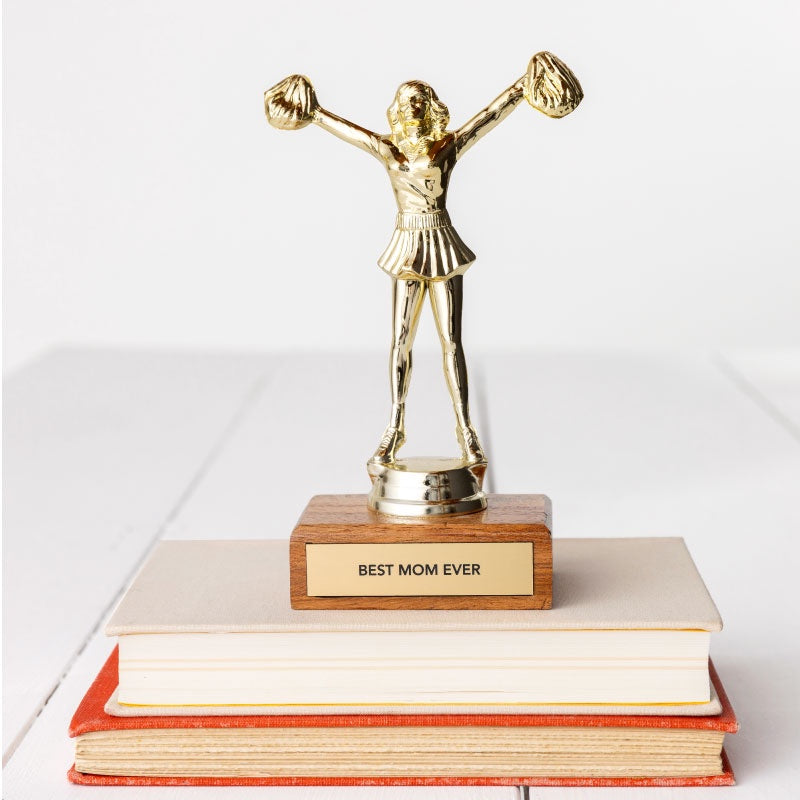 A JE Trophy depicting a woman with arms raised high, standing on a wooden base labeled &quot;BEST MOM EVER&quot;, placed atop a stack of books in Bungalow style on a white background. (Brand: Faire)