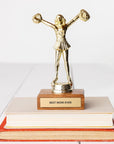 A JE Trophy depicting a woman with arms raised high, standing on a wooden base labeled "BEST MOM EVER", placed atop a stack of books in Bungalow style on a white background. (Brand: Faire)