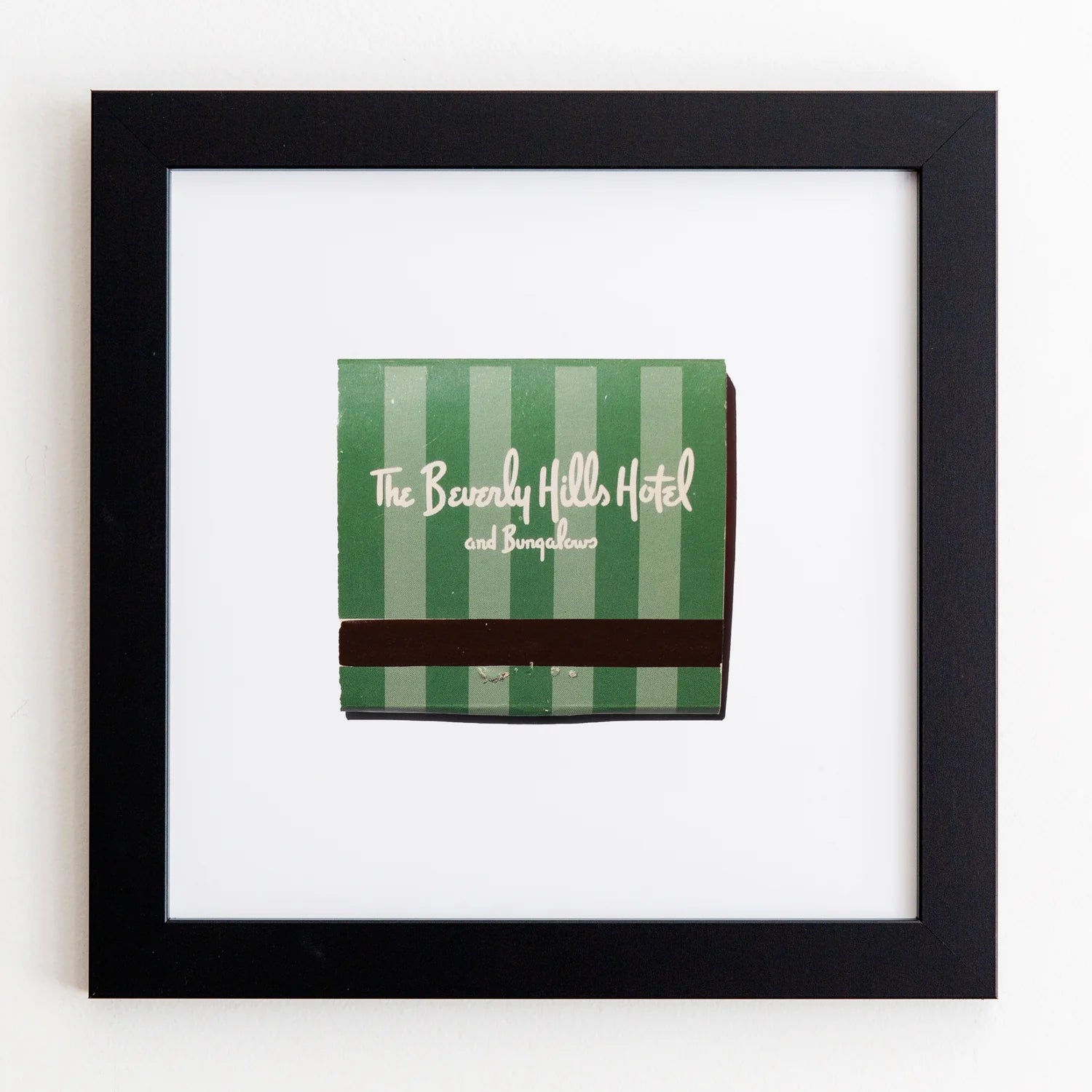 A framed matchbook from Match South, featuring a green and white striped cover with brown accents, mounted on a white background in an Art Square Black Frame.