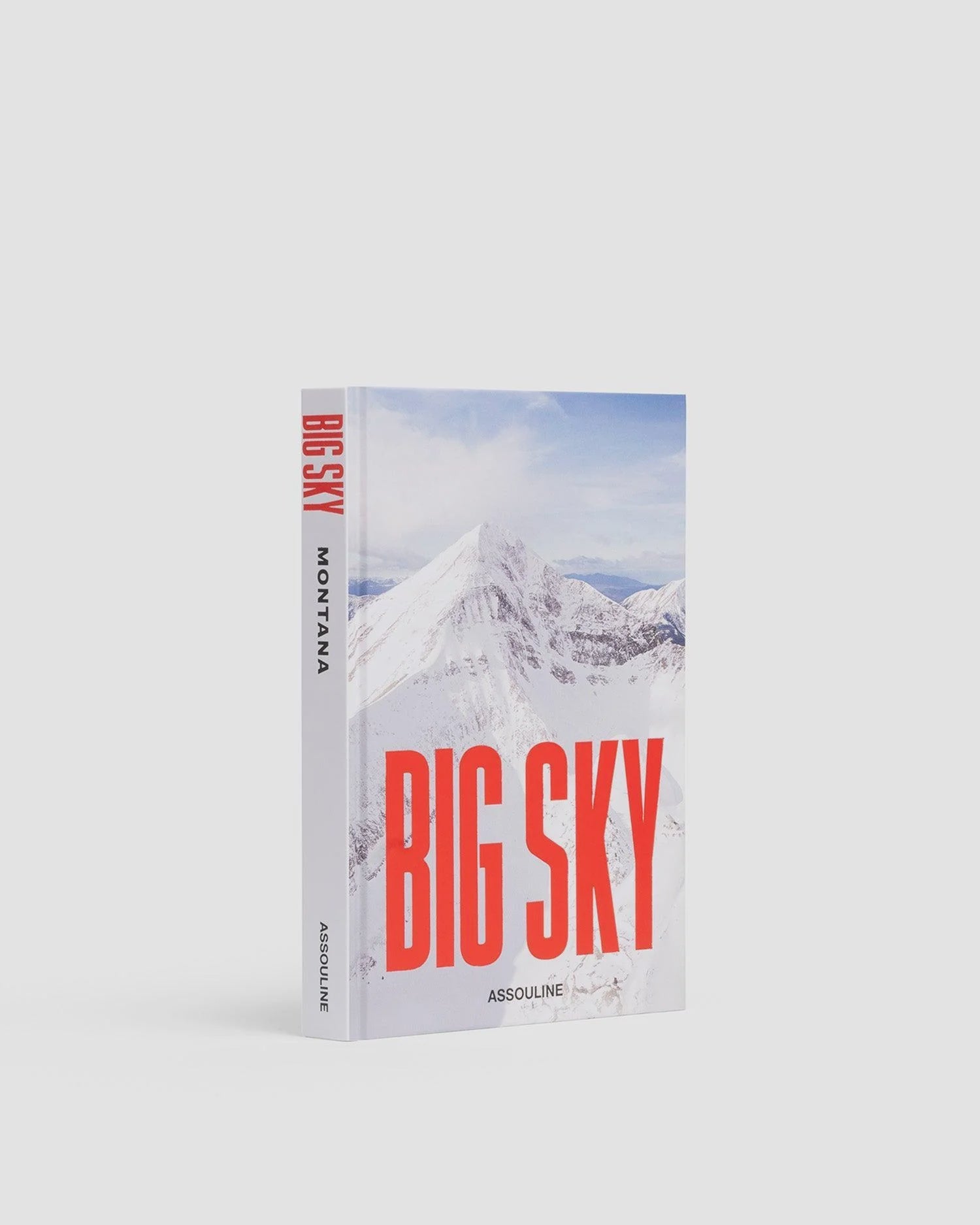 A hardcover book titled "Big Sky" by Assouline, ideal for outdoor enthusiasts. The cover showcases a stunning photo of snow-covered mountains, with the spine elegantly displaying "Big Sky Montana" and "Assouline" at the bottom. Set against a plain white background, it invites dreams of thrilling skiing adventures.