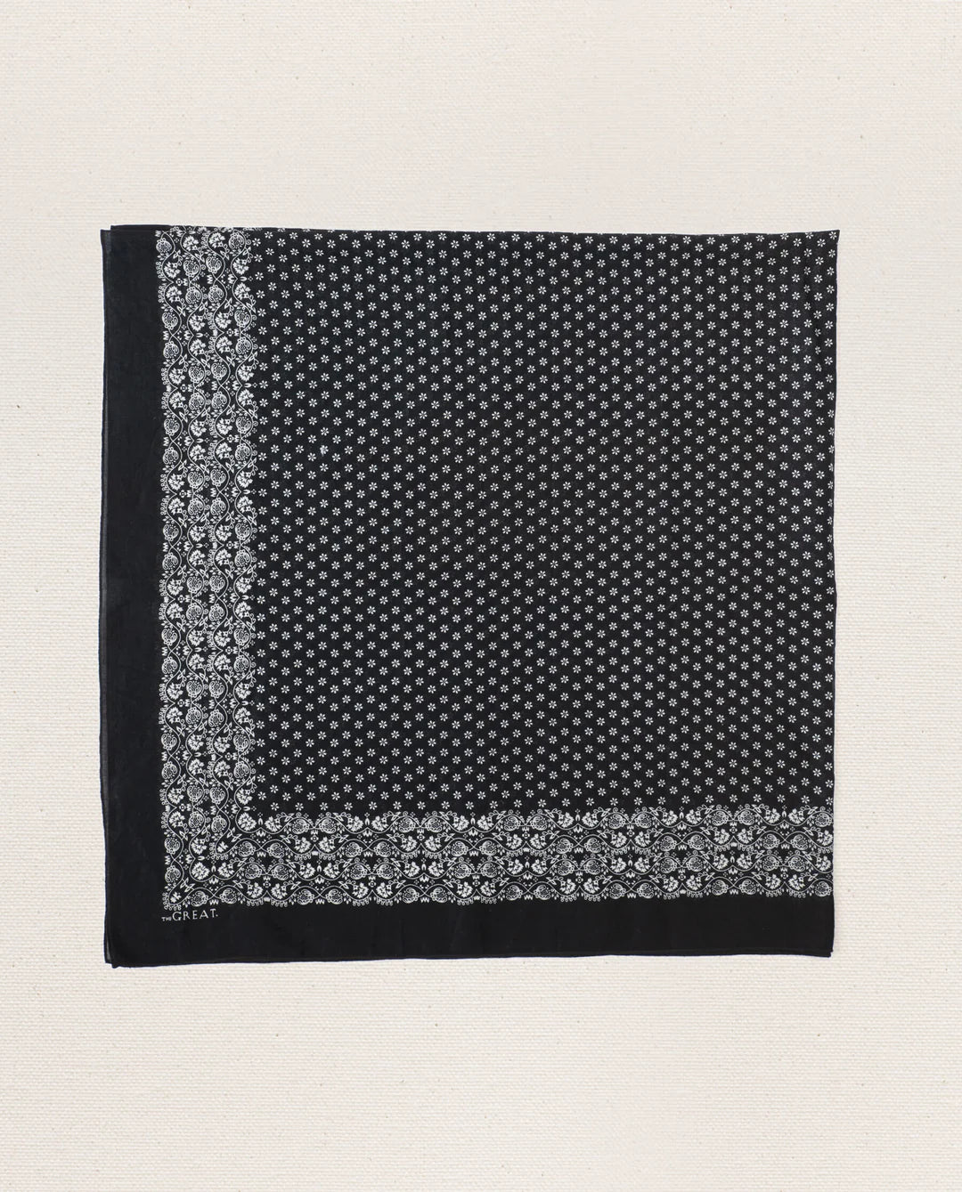 The Great Inc.'s The Big Bandana is a square black cotton scarf with a white floral border and dense white dots in the center, vintage-inspired and displayed flat on a neutral textured background.
