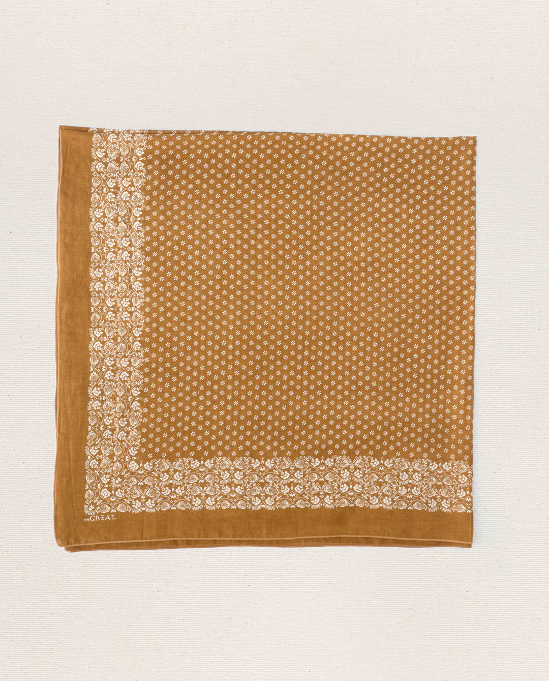 The Big Bandana by The Great Inc. is a square vintage cotton bandana with a brown background, white floral border, and small white floral patterns in the center, shown laid flat on a light textured surface.