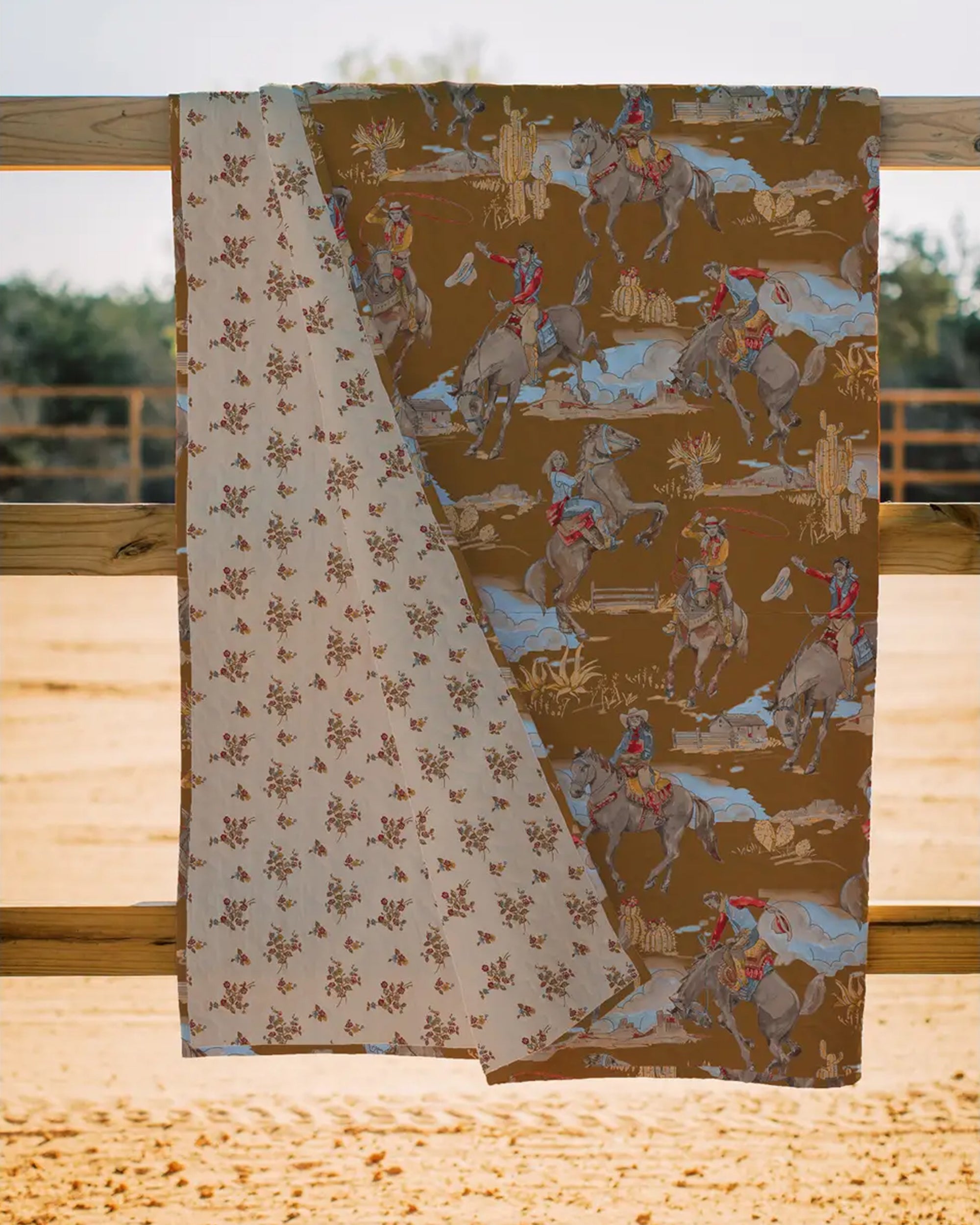 The Birdie Gold Quilt by Faire hangs over a wooden fence; one side displays cowboys on horseback against brown, while the reverse features red and brown florals on cream—a cozy, two-sided Texas-made quilt for tradition lovers.