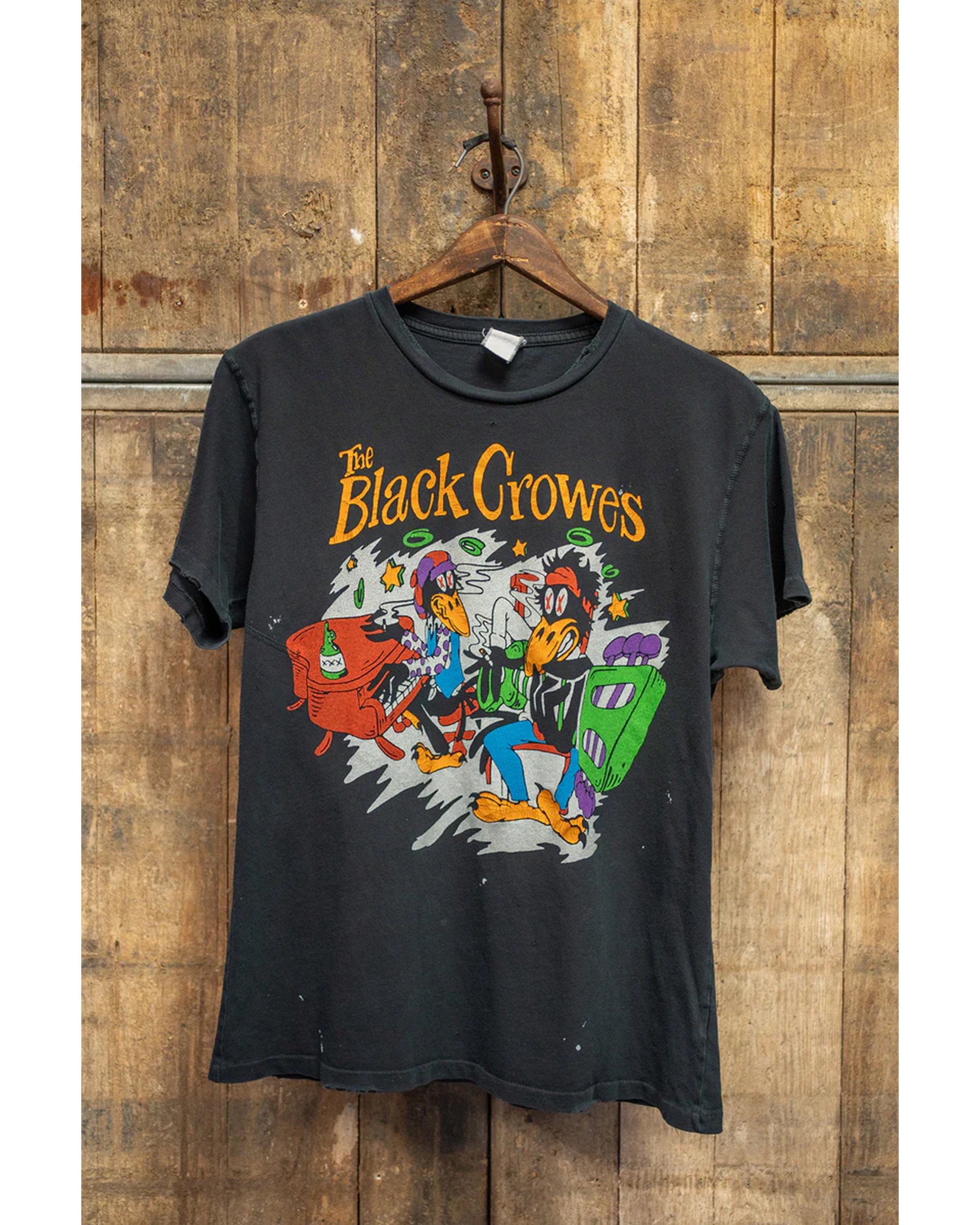 The Black Crowes Destroyed Tee Coal by Made Worn hangs on a wooden hanger against a rustic wall. This vintage unisex shirt features colorful cartoon crows playing instruments and "The Black Crowes" text on signature cotton.